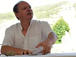 Assis à une table devant des notes, un homme en chemise blanche tourne la tête vers sa gauche.