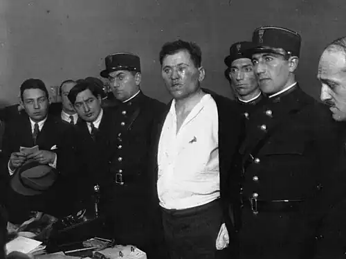 Photo en noir et blanc d’agents des forces de l’ordre encadrant un homme blessé au visage, l’air hagard, avec une chemise blanche entrouverte tâchée de sang