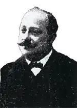  Portrait photo de mauvaise qualité d'un homme 50-60 ans, front dégarni et moustache, costume sombre et nœud papillon.