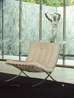 fauteuil blanc, armature en métal dans une pièce ; à l’arrière-plan, à l’extérieur de la vitre dans une cour fermée, une statue de femme nue.