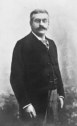 Photo en noir et blanc longueur trois-quarts d'un homme se tenant debout, avec les cheveux grisonnants, des sourcils et une moustache foncés, portant des lorgnons, un foulard, une chemise blanche, un veston et une redingote noirs, un pantalon clair.