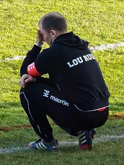 Un homme en survêtement sur le bord d'un terrain, accroupi.