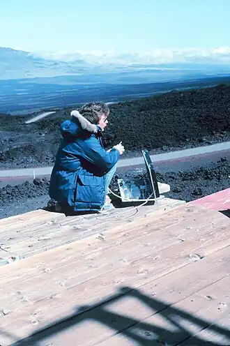 Un homme en veste bleue et avec des lunettes de soleil, est accroupi devant une mallette ouverte qui contient du matériel scientifique. Il se tient sur un bardage en bois, lui-même posé sur des roches noires. En arrière plan, la pente descendante du volcan et un panorama sur Hawaï.