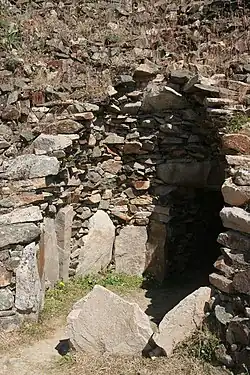 Vue de trois quarts d'une chambre en encorbellement éventrée. Des dalles sont plantées debout, en cercle, certaines incorporées dans la muraille. On aperçoit au fond à droite le débouché du couloir.