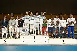 escrimeurs sur podium. 4 escrimeurs sont présents sur chacune des trois marches, un enfant figurant aussi sur la marche du milieu.