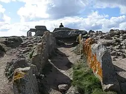 intérieur de l'allée couverte, en pente, vue depuis son entrée. La plupart des dalles de couvertures manquent. En arrière-plan, au sommet de l'éminence, on aperçoit le dolmen.