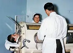 À la gauche, la tête d'une personne émerge d'un appareil. Deux personnes se tiennent debout de part et d'autre de l'appareil