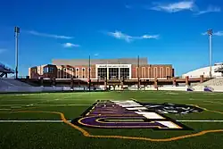 Photographie en contre-plongée d'un stade américain, vu depuis le terrain, en briques brunes sous le ciel bleu. Le symbole de l'équipe universitaire est présent au premier plan sur la pelouse.