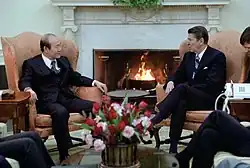 Photographie de deux hommes assis dans des fauteuils, disposés de part et d'autre d'une cheminée, face au photographe.