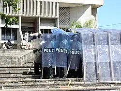 Plusieurs policiers armés en uniforme noir avec casques et boucliers. Protégés par un écran transparent en pvc sur un escalier devant un édifice.