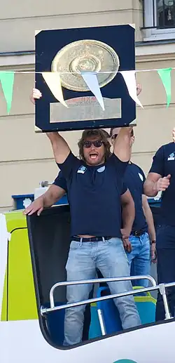 Homme en tenue de ville, de plein pied, tenant au-dessus une planche représentant le bouclier de Brennus.