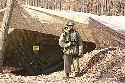 Soldat en uniforme dans une forêt. Son gilet tactique et son casque sont dans un motif de camouflage vert tandis que le reste de sa tenue est dans un motif camouflage beige.
