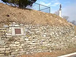Photographie d'un mur de pierre avec un talus de terre au-dessus. L'ensemble mesure environ trois mètres de haut et une petite statue au sommet d'un pilier blanc est situé au sommet.
