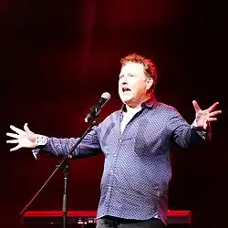 Photo d'un homme blanc aux cheveux courts bouclés. Il porte une chemise à motifs bleue et se tient devant un pied de micro. Il chante dans le micro, les bras écartés et la bouche grande ouverte.