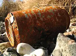 Photo en couleurs d’une canette de boisson rouillée et percée, abandonnée sur un terrain caillouteux.
