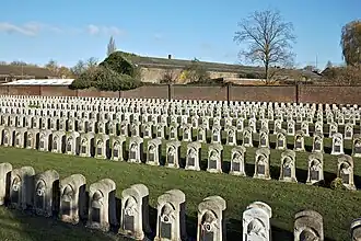 Photo de plusieurs rangées de stèles funéraires dans la pelouse. A l'arrière plan, on aperçoit un entrepôt derrière le mur d'enceinte en briques sombres.