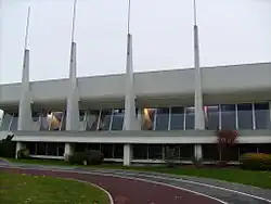 Photographie de la piscine, un bâtiment moderne en béton clair, aux lignes horizontales très marquées, éclairé par de larges baies vitrées. En façade, des sortes de contreforts s'élèvent du sol, allant en s'amenuisant, dépassant la toiture pour se terminer par de hauts mâts métalliques.