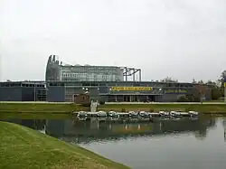 Photographie du Jardin du Monde, montrant un bâtiment bas incurvé, à structure de métal et panneaux pleins de couleur gris foncé, avec en son centre une serre enserrée dans un réseau métallique. Devant s’étale, entouré de pelouses, un plan d’eau avec un ponton et des petits bateaux à quai.