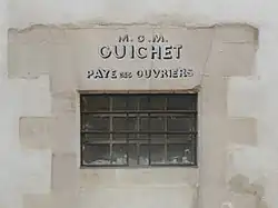 Fenêtre étroite percée dans un mur, sur lequel est inscrit «&nbsp;M.O.M. Guichet Paye des ouvriers&nbsp;».