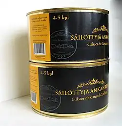 Deux boîtes identiques, à l’étiquette noire et orange avec un texte en lettre dorées annonçant des cuisses de canard confites, sont empilées exactement l’une au-dessus de l’autre. Les bords et le rétreint sont en métal doré.