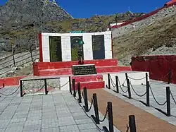 Mémorial en pierre rouge et blanc où sont fixées des plaques noires avec des listes de noms écrites en jaune, et en arrière-plan une crête rocheuse fortifiée.