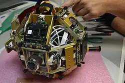 Un des satellites SPHERES sans sa coque en plastique. La structure en aluminium, le panneau de contrôle, les capteurs à ultrasons, propulseurs, la molette du régulateur de pression et le manomètre de pression sont visibles.
