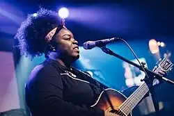 Une femme noire bouche ouverte devant un micro. Elle tient une guitare.