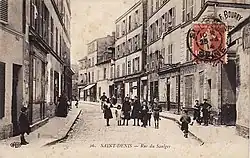 Carte postale sépia d'une rue au début des années 1900. Des enfants prennent la pose au milieu de la voie. Légende : « Saint-Denis, rue du Saulger ».