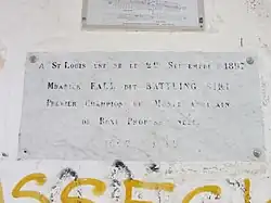 Est écrit en lettres capitales sur une plaque commémorative placée sur un mur : « A St Louis, est né le 22 septembre 1897 Mbarick Fall dit Battling Siki premier champion du monde africain de boxe professionnelle - 1897 – 1925 »