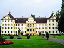 Vue d'un grand bâtiment à la façade blanche et jaune, devant lequel se trouve le buste d'un homme et un jardin.