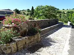 Fontaine en pierre surmntée de fleurs, sur une place de village pavée, plus loin le paysage est composé d'arbres et de prés verts.