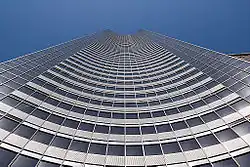 Photographie du bâtiment depuis le bas jusqu'au ciel. Il forme une sorte de gouttière ouverte concave.