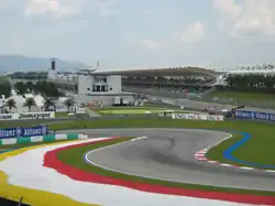 Piste goudronnée de Formule 1 au niveau d'une chicane, avec une tribune en arrière-plan.