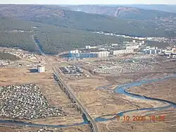 Photographie aérienne montrant une route passant au milieu au-dessus d'une rivière sinueuse et de ses berges marécageuses. Sur le côté gauche du pont se trouve un quartier de maisons tandis qu'un haut à droite se trouvent de grands ensembles soviétiques. Au fond la route s'enfonce dans la taïga et des collines se distinguent.