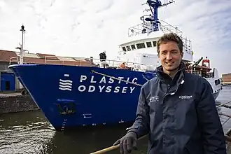 Simon Bernard pose devant le bateau Plastic Odyssey, mis à l'eau derrière lui.