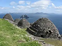 Photographie en couleurs de trois bâtiments en forme de dôme, appareillés de pierres grises et blanches, un pré s'étendant sur la gauche, une mer et des îles visibles sur la droite.