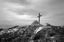 Photographie en noir en blanc d'une croix sommitale en bois et d'un cairn avec un ciel nuageux.