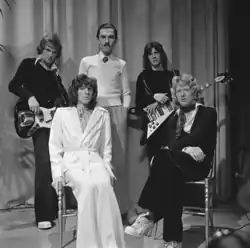 Photo en noir et blanc de cinq jeunes hommes blancs. Trois sont debout à l'arrière, dont deux équipés de guitares. Le troisième, au milieu, se tient les bras le long du corps. Deux sont assis à l'avant, celui à gauche est entièrement habillé en blanc.