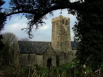 Photo d'un bâtiment allongé aux murs gris et au toit en ardoises, avec une tour carrée trapue en son centre. La silhouette d'un arbre au premier plan encadre le sujet