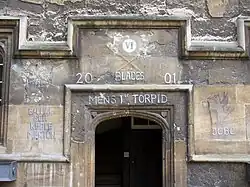 Une arche de pierre, sans porte ; au-dessus, les chiffres romains "VI" dans un cercle blanc et, à la craie, deux avirons avec des pales vertes, les mots "Blades", "2001", "Men's 1st Torpid" ; à gauche, l'inscription "Balliol SEH Keble Merton", à droite, un dragon debout tenant un drapeau au-dessus des initiales «JCBC"