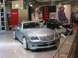 Une voiture grise 2 portes vue de l'avant droit exposée dans un salon aux côtés d'autres automobiles. Une femme s'appuie sur la portière conducteur de la voiture.