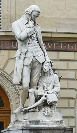 Statue représentant Valentin Haüy, réalisée par Badiou de la Tronchère. Elle est située à l'Institut des jeunes aveugles à Paris. Valentin Haüy semble pensif, la main droite sur la tempe, la main gauche posée affectueusement sur un jeune aveugle assis à ses pieds, qui est en train de lire un livre en braille.