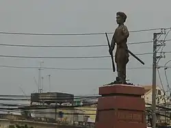 Vue de loin d'une statue en ciment montrant un homme moustachu tenant un fusil dans une main et un bâton dans l'autre, portant des vêtements traditionnels. Il est debout sur un piédestal de pierre polie.