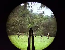 photographie en couleur montrant un pré et une forêt vus à travers un viseur circulaire avec une barre verticale au centre