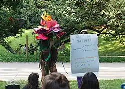 Deux personnes debout dans un parc devant un groupe assis. Une performeuse, porte un costume de plante : elle est a une grande fleur rose, jaune et violette en tissu sur la tête ainsi qu'une robe en crochet verte et marron et tient un micro. L'autre personne se devine derrière une grande affiche, sur laquelle est écrit "Pas d'ordre naturel. Anomalies → adaptation et non pas effondrement. La diversité transpédégouine est un stade avancé et créatif de l'évolution"