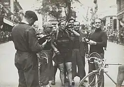 Photographie en noir et blanc d'un cycliste buvant à sa gourde et entouré par plusieurs hommes après l'arrivée de la course.