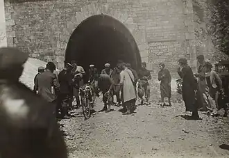 Photographie en noir et blanc d'un cycliste démontant sa roue à la sortie d'un tunnel, sur une piste caillouteuse, entouré par plusieurs hommes.