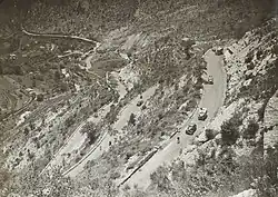 Vue plongeante sur des cyclistes suivis par plusieurs véhicules sur une route de montagne.