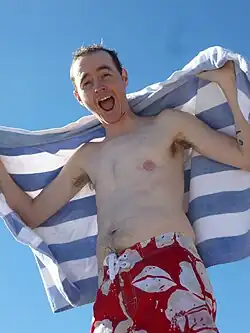 Photographie d'un homme trans torse nu et souriant, dont le développement mammaire est réduit au minimum et dont la poitrine ne laisse apparaître aucune cicatrice.