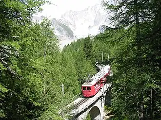 Photographie en couleurs du train en contrebas, des arbres de chaque côté, la montagne au fond